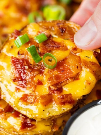 A hand holding a crispy slice of potato topped with melted cheese, bacon, and green onions.