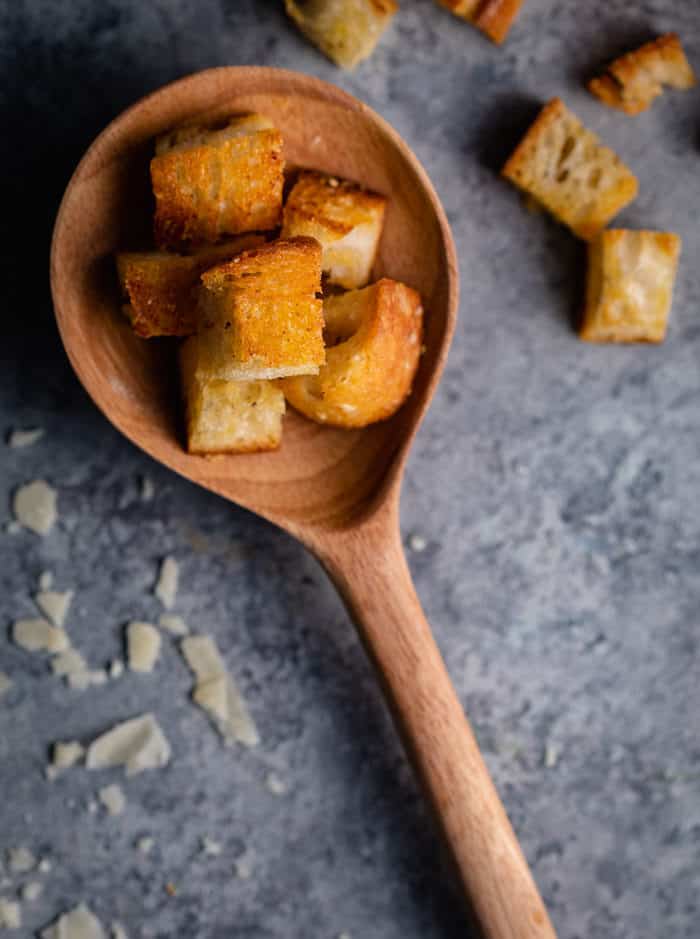wooden spoon holding homemade croutons on a shadowy surface sprinkled with grated Parmesan.