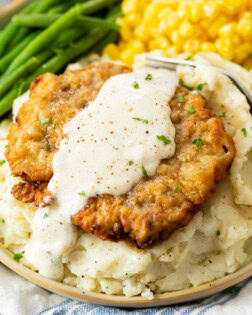 Crispy Chicken Fried Steak on a bed of mashed potatoes with white gravy on top and vegetables in the back.