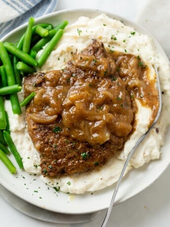 A pile of mashed potatoes topped with cubed steak with brown gravy and onions with green beans on the side.
