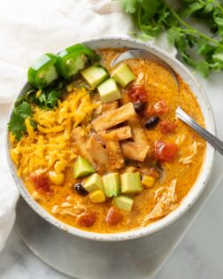 Overhead view of a white bowl with creamy chicken tortilla soup and garnishes like crispy tortillas, avocado, and shredded cheese.
