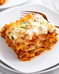 A white plate with Lasagna on it with a fork in the background.