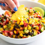 A hand dipping a tortilla chip into a white bowl of Mango Salsa.