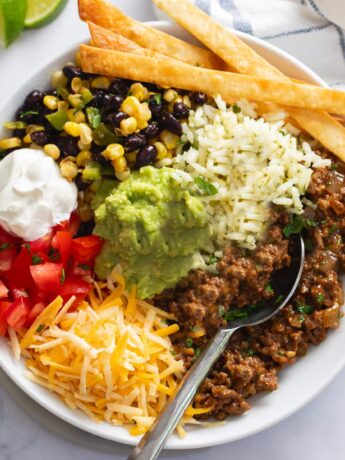 Burrito Bowls filled with rice, seasoned ground beef, corn, black beans, tomatoes, sour cream, and fried tortilla strips.