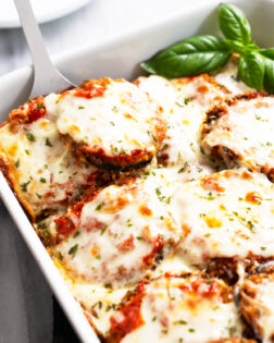 Eggplant Parmesan in a white casserole dish with a spatula scooping it up and fresh basil on the side.