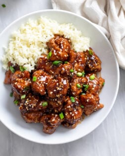 Crispy Sesame Chicken in a white bowl with rice on the side and green onions on top.