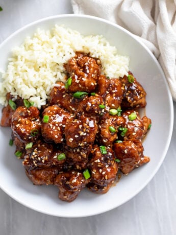 Crispy Sesame Chicken in a white bowl with rice on the side and green onions on top.
