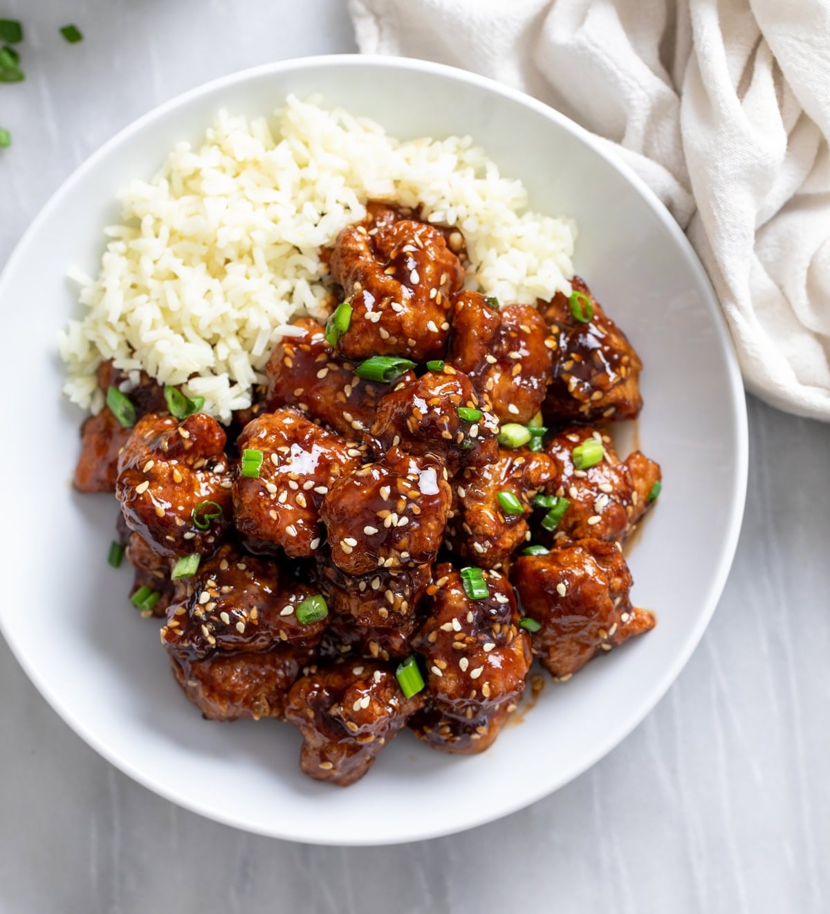 Crispy Sesame Chicken in a white bowl with rice on the side and green onions on top.