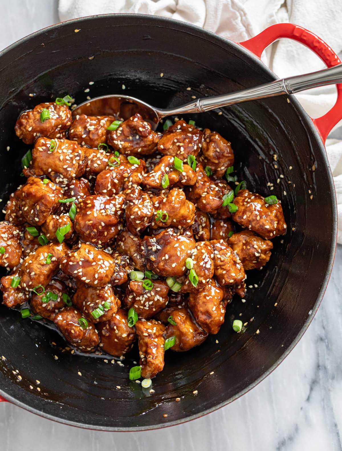 Sesame Chicken in a wok with sesame seeds and green onions on top and a spoon on the side.