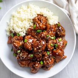 Sesame Chicken in a glossy sauce with rice on the side and sesame seeds and green onions on top.
