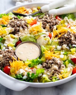 Side view of Big Mac Salad in a white serving bowl with homemade big mac dressing, lettuce, ground beef, onions, pickles, cheese, and tomatoes.