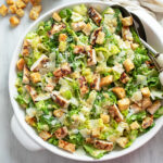 Chicken Caesar Salad in a white bowl with romaine lettuce, grilled chicken parmesan cheese, homemade dressing, and homemade croutons.