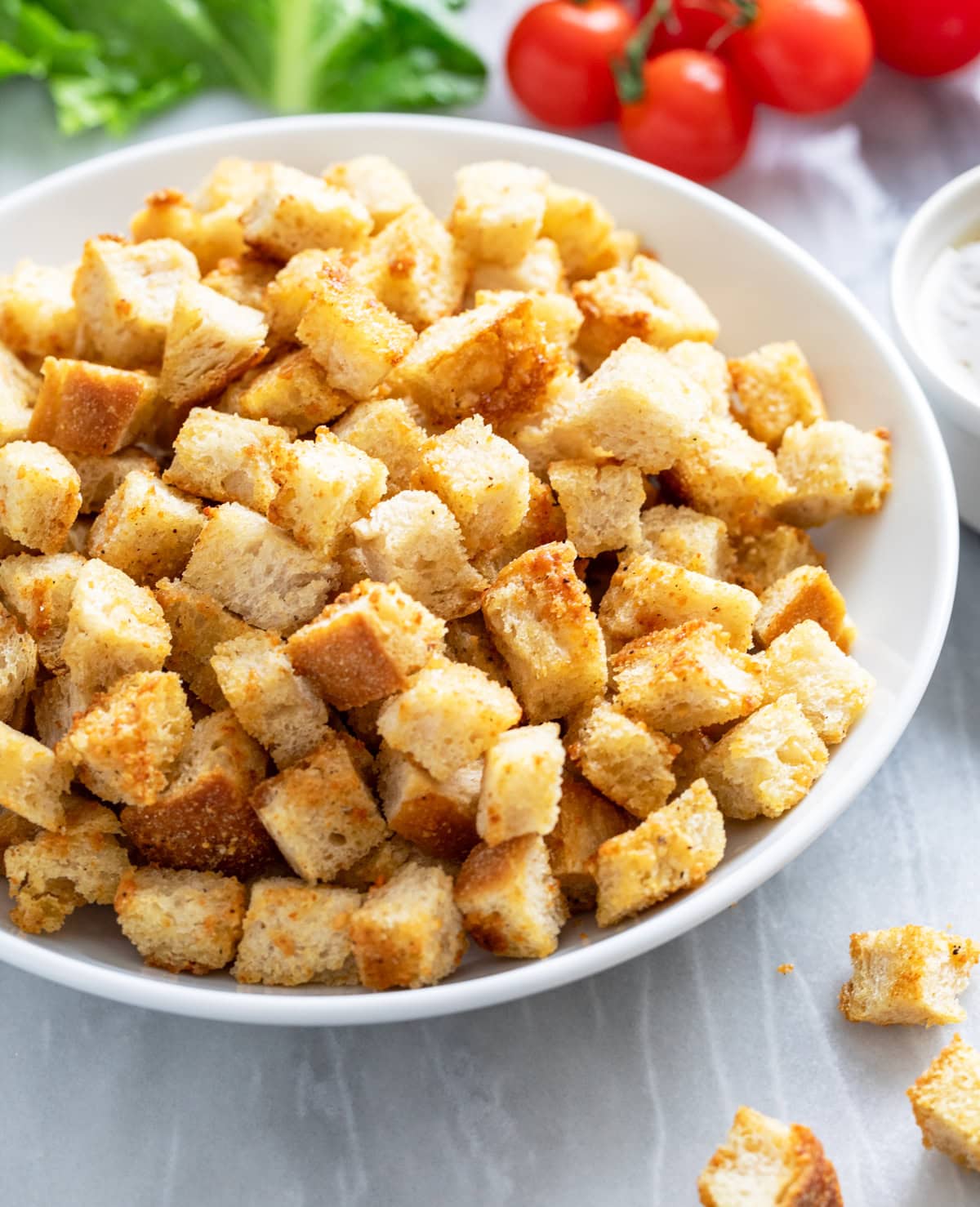 Side view of homemade croutons in a white bowl with tomatoes and lettuce in the background.