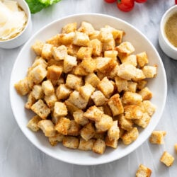 Homemade Croutons in a white bowl with sides of dressing, parmesan, lettuce, and tomatoes.