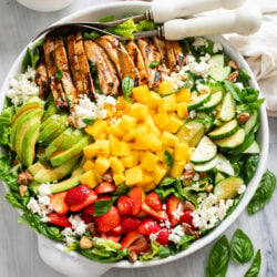 Overhead view of Mango Salad in a white bowl with romaine, spinach, grilled chicken, avocado, strawberries, feta, mango, and homemade dressing.