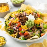 Taco Salad in a big white bowl topped with seasoned ground beef, guacamole, sour cream, tomatoes, corn, black beans, onions, cheese, and more.
