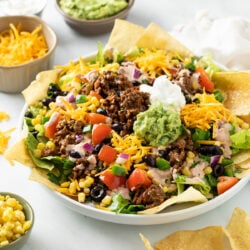 Taco Salad in a big white bowl topped with seasoned ground beef, guacamole, sour cream, tomatoes, corn, black beans, onions, cheese, and more.