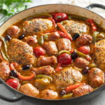 Side view of Chicken Scarpariello in a braiser with pan sauce, sausage, chicken thighs, peppers, and cherry peppers.